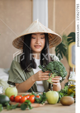A young Asian woman wearing a conical hat prepares fresh broccoli and other vegetables for a healthy meal A young Asian woman wearing a conical hat prepares fresh broccoli and other vegetables for a healthy meal 135627765