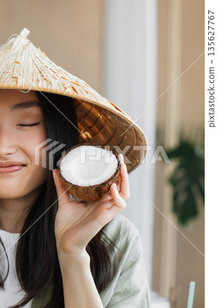 A young woman wearing a traditional conical hat holds a halved coconut near her face with a gentle smile A young woman wearing a traditional conical hat holds a halved coconut near her face with a gentle smile 135627767
