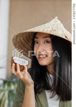 A young Asian woman wearing a conical hat holds a halved coconut, smiling at the camera A young Asian woman wearing a conical hat holds a halved coconut, smiling at the camera 135627768