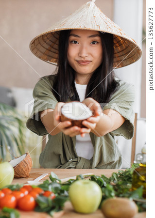 A young Asian woman wearing a conical hat offers a halved coconut with white flesh to the camera while surrounded by fresh ingredients on a wooden table 135627773