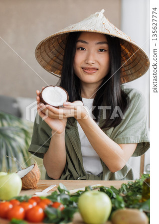 A young Asian woman wearing a conical hat holds a halved coconut while surrounded by fresh produce on a wooden table, suggesting healthy eating or cooking A young Asian woman wearing a conical hat holds a halved coconut while surrounded by fresh produce on a wooden table, suggesting healthy eating or cooking 135627774