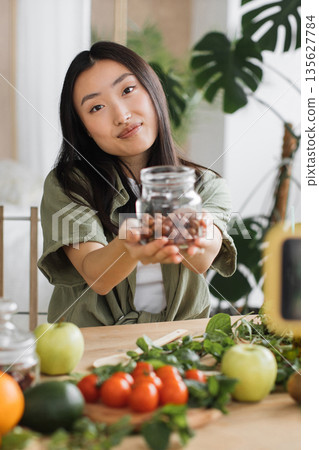 A young Asian woman smiles while presenting a jar filled with hazelnuts, surrounded by fresh fruits and vegetables on a wooden table, suggesting healthy eating and organic produce 135627784