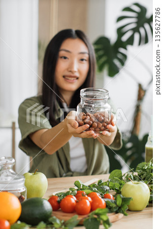 A young woman with dark hair smiles as she holds up a glass jar filled with nuts, surrounded by fresh fruits and vegetables on a wooden table A young woman with dark hair smiles as she holds up a glass jar filled with nuts, surrounded by fresh fruits and vegetables on a wooden table 135627786