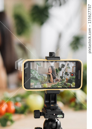 A young woman films herself preparing a healthy meal with fresh produce and plants in the background, showcasing a lifestyle vlog 135627787