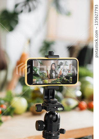 A young woman records a healthy food vlog on her smartphone, surrounded by fresh fruits and vegetables, showcasing a vibrant lifestyle A young woman records a healthy food vlog on her smartphone, surrounded by fresh fruits and vegetables, showcasing a vibrant lifestyle 135627793