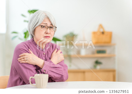 一位頭髮花白的老婦人在家裡的客廳或餐廳裡瑟瑟發抖（衣著單薄，怕冷，免疫力低下，發冷）。 135627951