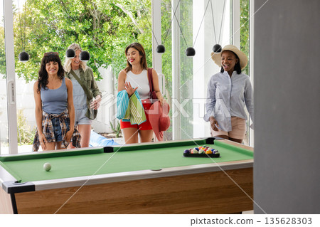 Group of women entering room with pool table, carrying beach towels and hats 135628303