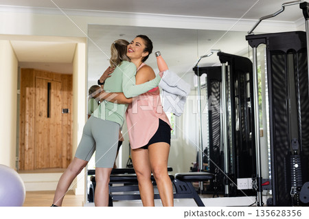 Women hugging in gym after workout, holding water bottle and towel, smiling Women hugging in gym after workout, holding water bottle and towel, smiling 135628356