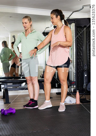 Women exercising together in gym, discussing workout routine near exercise equipment Women exercising together in gym, discussing workout routine near exercise equipment 135628376