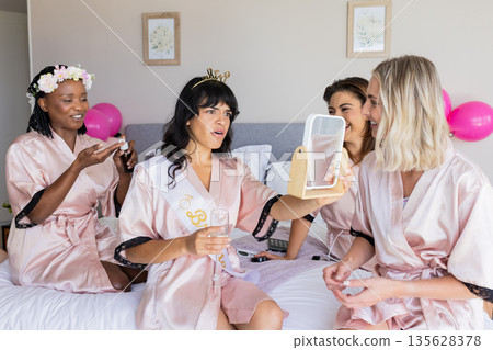 Women in robes enjoying bachelorette party, laughing and applying makeup on bed Women in robes enjoying bachelorette party, laughing and applying makeup on bed 135628378