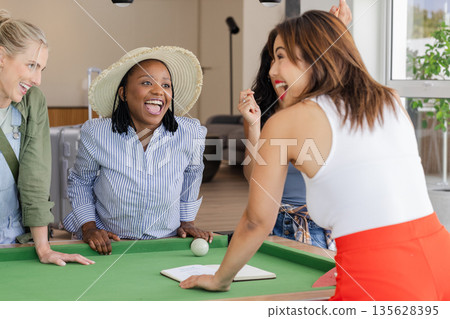 Women laughing and enjoying lively conversation around pool table indoors 135628395