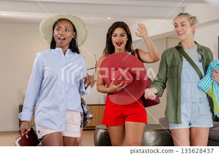 Three women joyfully entering hotel lobby, holding hats and beachwear Three women joyfully entering hotel lobby, holding hats and beachwear 135628437