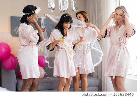 Women in robes celebrating bachelorette party, helping bride-to-be with veil 135628446