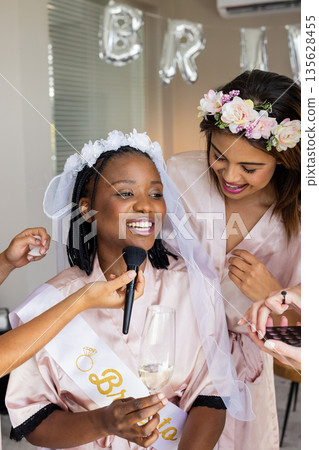 Celebrating bridal shower, African American bride smiling while friends apply makeup Celebrating bridal shower, African American bride smiling while friends apply makeup 135628455