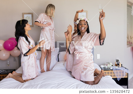 Women in robes celebrating on bed with champagne and balloons, enjoying party 135628472
