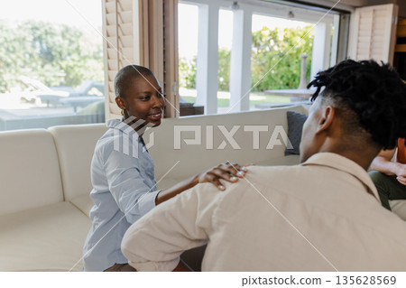 Smiling woman sitting on sofa, placing hand on friend's shoulder, offering support Smiling woman sitting on sofa, placing hand on friend's shoulder, offering support 135628569