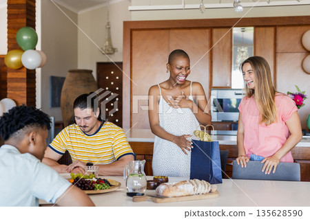 Friends celebrating at home, sharing gifts and laughter around dining table 135628590