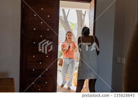 Friends greeting each other warmly at doorway, sharing excitement and joy Friends greeting each other warmly at doorway, sharing excitement and joy 135628643