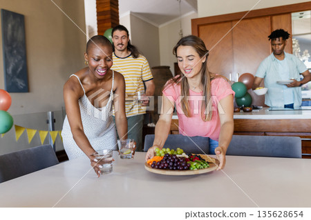 Friends setting table with fruit platter and drinks, preparing for indoor gathering 135628654