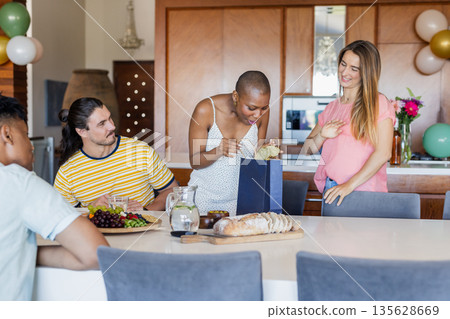 Friends celebrating at home, woman opening gift bag with excitement and joy Friends celebrating at home, woman opening gift bag with excitement and joy 135628669