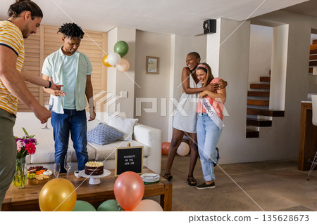 Friends celebrating engagement at home, hugging and smiling with cake and balloons Friends celebrating engagement at home, hugging and smiling with cake and balloons 135628673