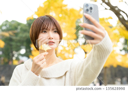 A woman with short hair taking a selfie with her smartphone against the backdrop of a ginkgo tree A woman with short hair taking a selfie with her smartphone against the backdrop of a ginkgo tree 135628998