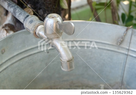 Outdoor silver faucet attached to metal basin delivering water flow in garden setting with worn pipe connection for utility washing purpose. Outdoor silver faucet attached to metal basin delivering water flow in garden setting with worn pipe connection for utility washing purpose. 135629362