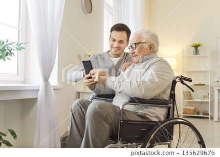 Nurse Helping Elderly Patient In Wheelchair With Smartphone And Entertainment 135629676