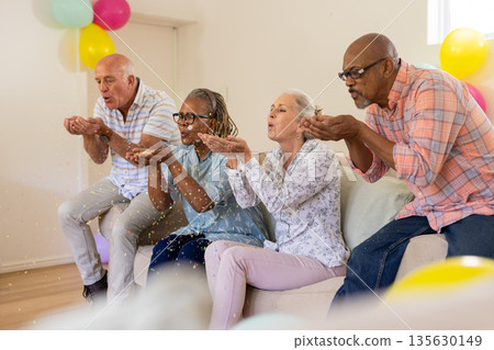 Senior friends celebrating together, blowing confetti in living room, enjoying party Senior friends celebrating together, blowing confetti in living room, enjoying party 135630149