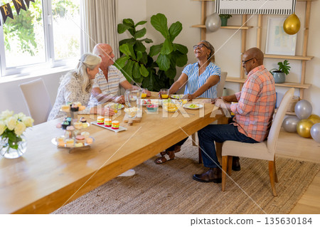 Senior friends laughing and enjoying snacks together at home around dining table 135630184