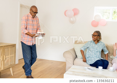 Senior man carrying birthday cake while friends smiling and celebrating at home Senior man carrying birthday cake while friends smiling and celebrating at home 135630211