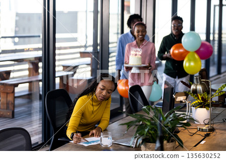 Colleagues surprising woman with cake and balloons in modern office setting 135630252