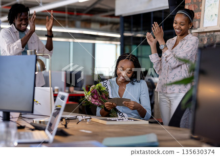 African American woman reading card with flowers, colleagues applauding in office African American woman reading card with flowers, colleagues applauding in office 135630374