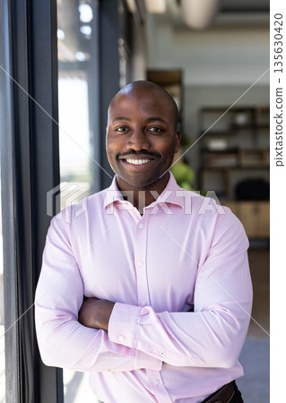 Confident businessman in pink shirt standing by office window, smiling warmly 135630420