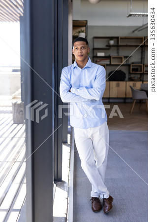 Confident businessman in blue shirt standing by office window, looking thoughtful 135630434
