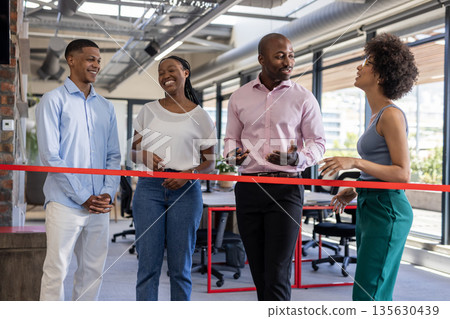Colleagues celebrating office opening, smiling and chatting near red ribbon indoors 135630439