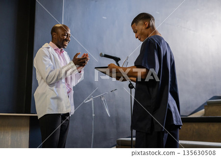 Man in white coat clapping while another man reads from clipboard on stage 135630508