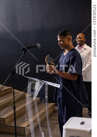 Young man in scrubs holding award at podium, smiling with colleague nearby Young man in scrubs holding award at podium, smiling with colleague nearby 135630522