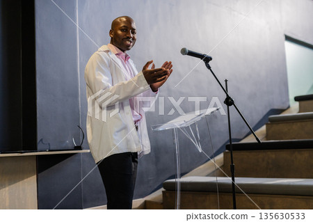 Businessman in white jacket clapping while speaking at podium in conference hall Businessman in white jacket clapping while speaking at podium in conference hall 135630533