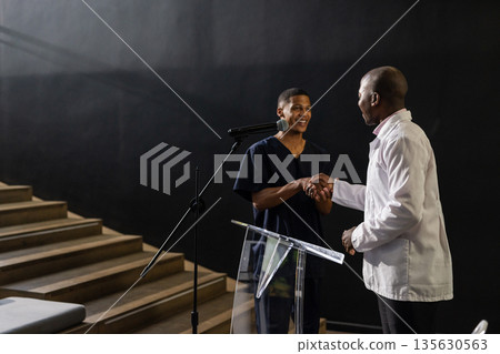 Two men shaking hands on stage, smiling and engaging in conversation Two men shaking hands on stage, smiling and engaging in conversation 135630563