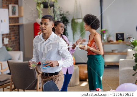 Holding drinks at office party, African American man with colleagues chatting nearby 135630631