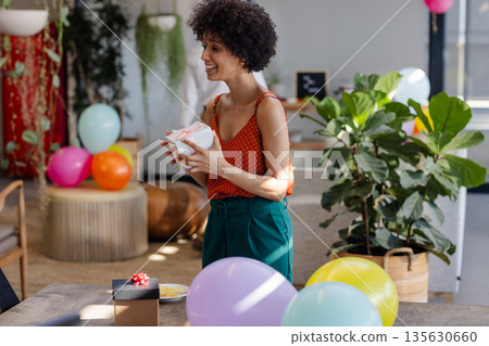 Smiling woman holding gift at office party with colorful balloons around 135630660