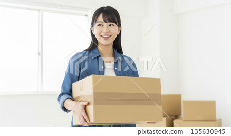 一位身穿牛仔襯衫的日本女子麵帶微笑，一邊回頭張望，一邊抱著搬家用的紙箱。 135630945
