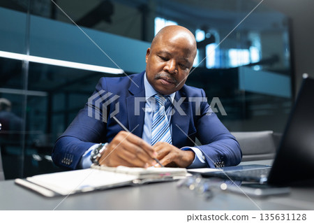 Black mature businessman writing notes at desk in modern office Black mature businessman writing notes at desk in modern office 135631128