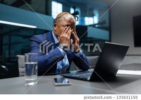 Exhausted mature black businessman rubbing eyes while sitting at desk in office Exhausted mature black businessman rubbing eyes while sitting at desk in office 135631150