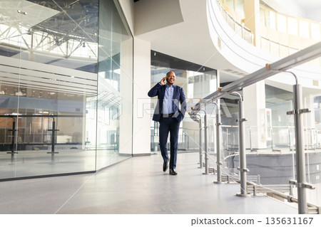 Black mature businessman walking confidently through office corridor while talking on smartphone Black mature businessman walking confidently through office corridor while talking on smartphone 135631167