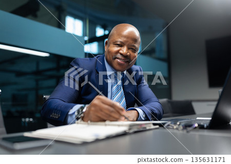 Black mature businessman smiling while writing notes at office desk 135631171