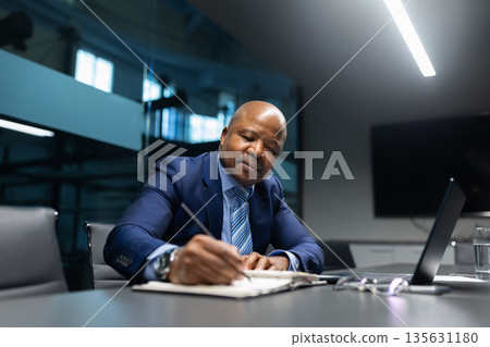 Black mature businessman writing at notepad while sitting at table in office 135631180