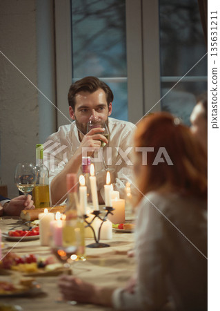Man drinking white wine at candlelit dinner table with friends in background. 135631211