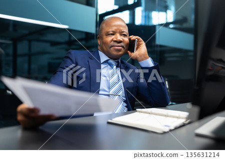 Confident black mature businessman holding documents while speaking on phone in office Confident black mature businessman holding documents while speaking on phone in office 135631214
